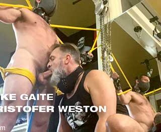 Mike Gaite stands naked, bound in bright yellow rope. A chain is attached to a leather collar around his neck which holds a metal hook, set deep in his ass. Kristofer Weston sits back stroking his cock through his shorts...