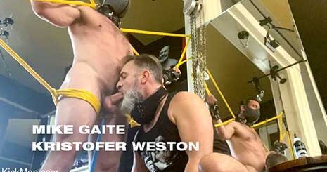 Mike Gaite stands naked, bound in bright yellow rope. A chain is attached to a leather collar around his neck which holds a metal hook, set deep in his ass. Kristofer Weston sits back stroking his cock through his shorts...